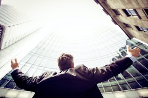 Business man from behind looking up at buildings in daylight with hands happily raised. 